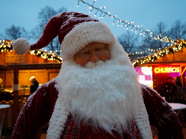 Sülzer Weihnachtsdorf Weihnachtsmann mit weißem Bart und roter Mütze, umgeben von festlich beleuchteten Ständen.