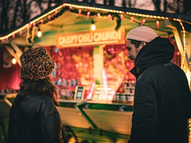 Two people in winter clothing standing before a brightly lit outdoor market stall at dusk.
