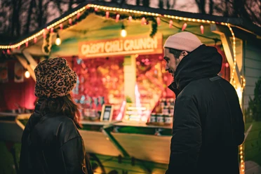 Zwei Personen vor beleuchtetem Weihnachtsmarktstand mit warmen Lichtern in der Dämmerung.