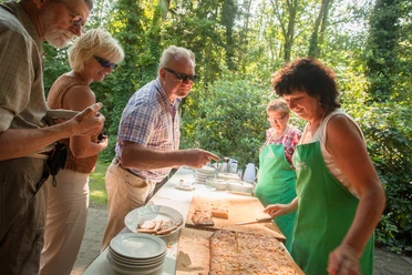 bispingen-butterkuchen-4311 Menschen genießen im Freien frischen Butterkuchen, serviert von Frauen mit grünen Schürzen.