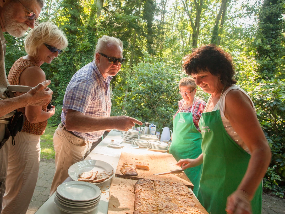 bispingen-butterkuchen-4311 Menschen genießen im Freien frischen Butterkuchen, serviert von Frauen mit grünen Schürzen.