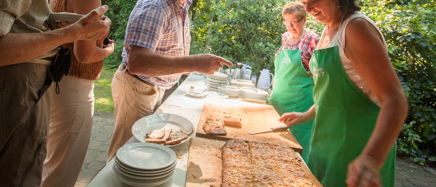 bispingen-butterkuchen-4311 Menschen genießen im Freien frischen Butterkuchen, serviert von Frauen mit grünen Schürzen.