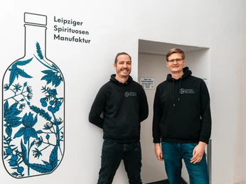 Gründer der Leipziger Spirituosen Manufaktur - Regionale Produkte aus Leipzig Die Gründer der Leipziger Spirituosen Manufaktur stehen vor einer Wandbemalung mit kreativem Flaschendesign