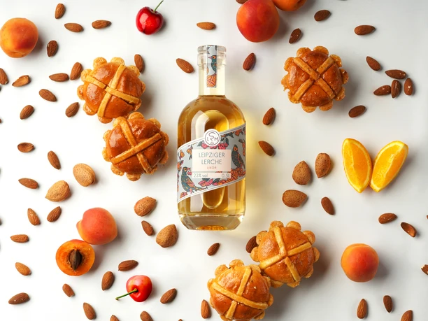 Zdjęcie produktowe butelki likieru Leipziger Lerchen na białym tle, otoczonej dekoracyjnymi składnikami, takimi jak prawdziwy likier Leipziger Lerche, morela i cynamon.