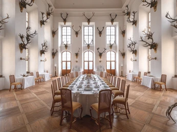 Geweihsammlung im Speisesaal.jpg Ein Speisesaal mit Kandelabern und Geweihen an weißen Wänden und einem gedeckten Tisch.