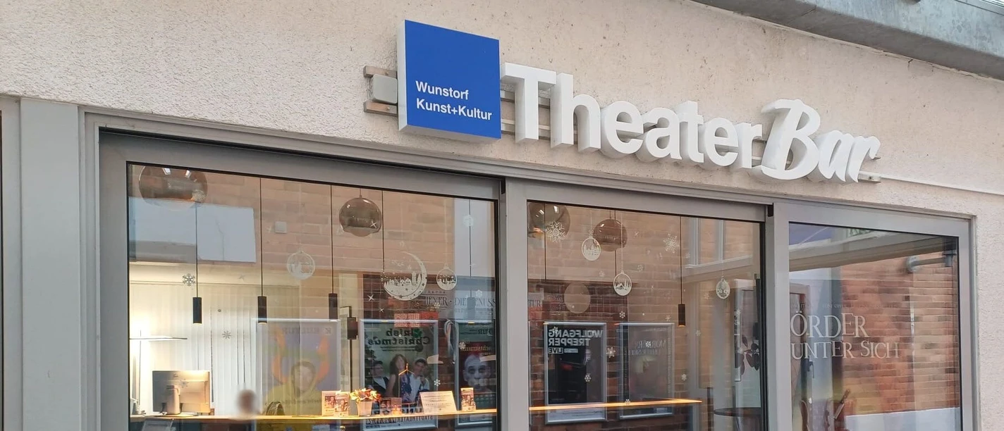 TheaterBar_Aussenansicht Eingang der TheaterBar in Wunstorf mit Glasfront, Plakaten und einem blauen Schild über der Tür.