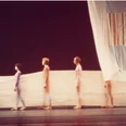 Stage design for Cunningham's choreography Travelogue Vor einem dunklen Bühnenhintergrund stehen in einer Reihe mehrere Menschen eng zusammen, eingehüllt in durchscheinende, farbig leuchtende Stoffbahnen, die gemeinsam ein ästhetisches Bild voller Anmut und Bewegung formen.Against a dark stage backdrop, several people stand close together in a row, wrapped in translucent, brightly colored lengths of fabric, which together form an aesthetic image full of grace and movement.