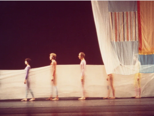 Stage design for Cunningham's choreography Travelogue Vor einem dunklen Bühnenhintergrund stehen in einer Reihe mehrere Menschen eng zusammen, eingehüllt in durchscheinende, farbig leuchtende Stoffbahnen, die gemeinsam ein ästhetisches Bild voller Anmut und Bewegung formen.Against a dark stage backdrop, several people stand close together in a row, wrapped in translucent, brightly colored lengths of fabric, which together form an aesthetic image full of grace and movement.