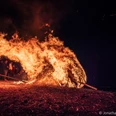 Ein großer, lodernder Feuerschwall neigt sich nach rechts, umgeben von einer Gruppe von Menschen bei Nacht.