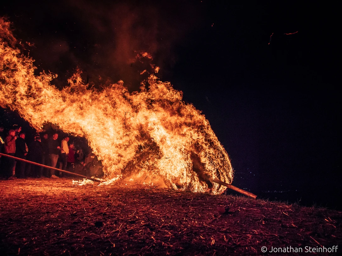 286a.jpeg Ein großer, lodernder Feuerschwall neigt sich nach rechts, umgeben von einer Gruppe von Menschen bei Nacht.