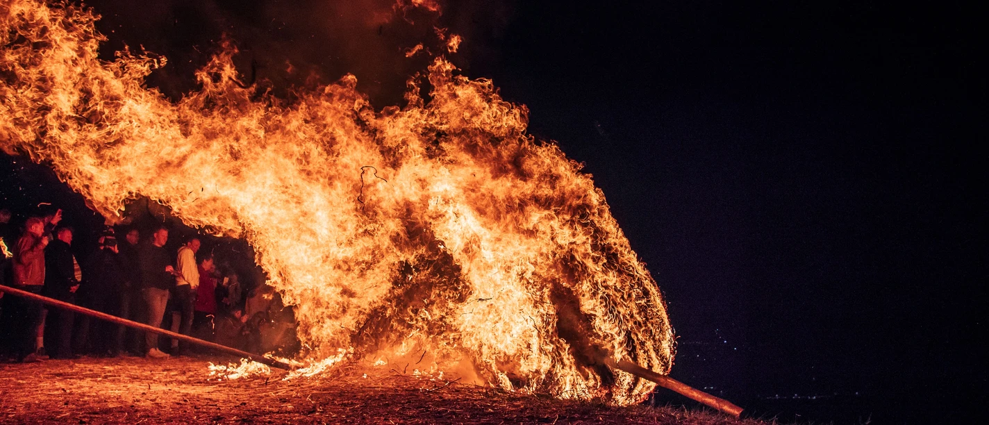 286a.jpeg Ein großer, lodernder Feuerschwall neigt sich nach rechts, umgeben von einer Gruppe von Menschen bei Nacht.
