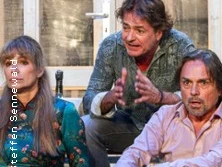 Drei Personen auf Theaterbühne, engagiert in lebhaftem Dialog vor rustikaler Dekor-Kulisse.