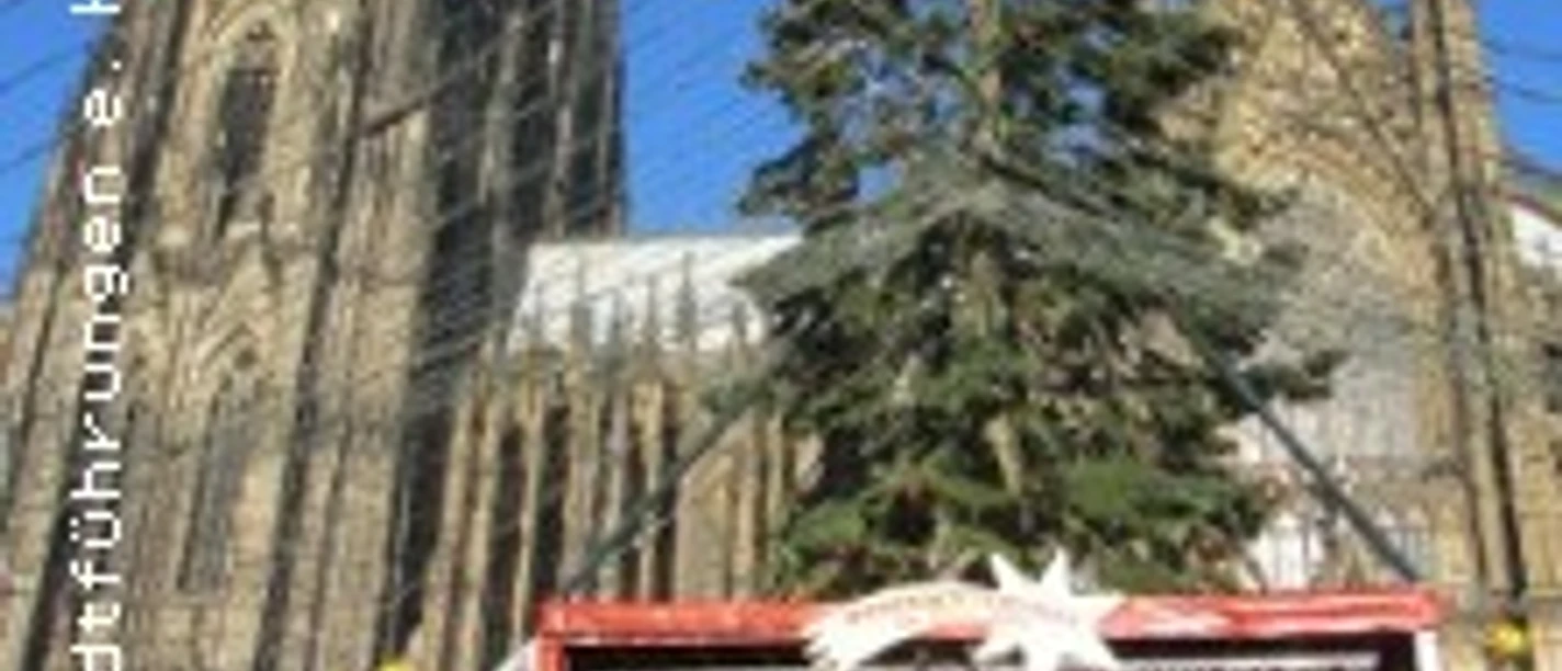 Kölner Dom im Hintergrund, ein geschmückter Weihnachtsbaum davor, festliche Bühne unter blauem Himmel.