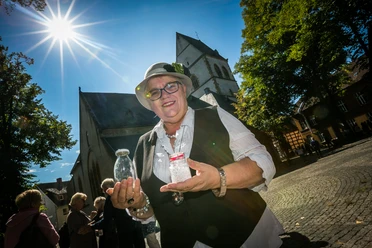 haller_herbst_20.jpg Ältere Frau in stilvoller Kleidung, lächelt vor historischer Kirche im Sonnenlicht, mit Glas in der Hand.