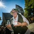 haller_herbst_20.jpg Ältere Frau in stilvoller Kleidung, lächelt vor historischer Kirche im Sonnenlicht, mit Glas in der Hand.