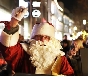 Nikolaus in rotem Mantel mit weißem Bart winkt in stimmungsvoll beleuchteter Einkaufsstraße.