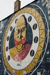 Mural Ein großflächiges Wandgemälde zeigt einen älteren Mann mit Brille, dessen Mund von einem roten Tuch bedeckt ist, eingerahmt in einem stilisierten Musterkreis.