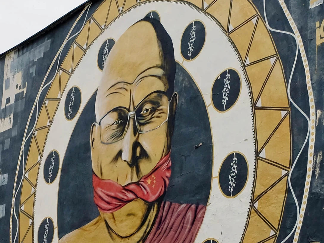 Mural Ein großflächiges Wandgemälde zeigt einen älteren Mann mit Brille, dessen Mund von einem roten Tuch bedeckt ist, eingerahmt in einem stilisierten Musterkreis.A large mural shows an elderly man with glasses, his mouth covered by a red cloth, framed in a stylized patterned circle.