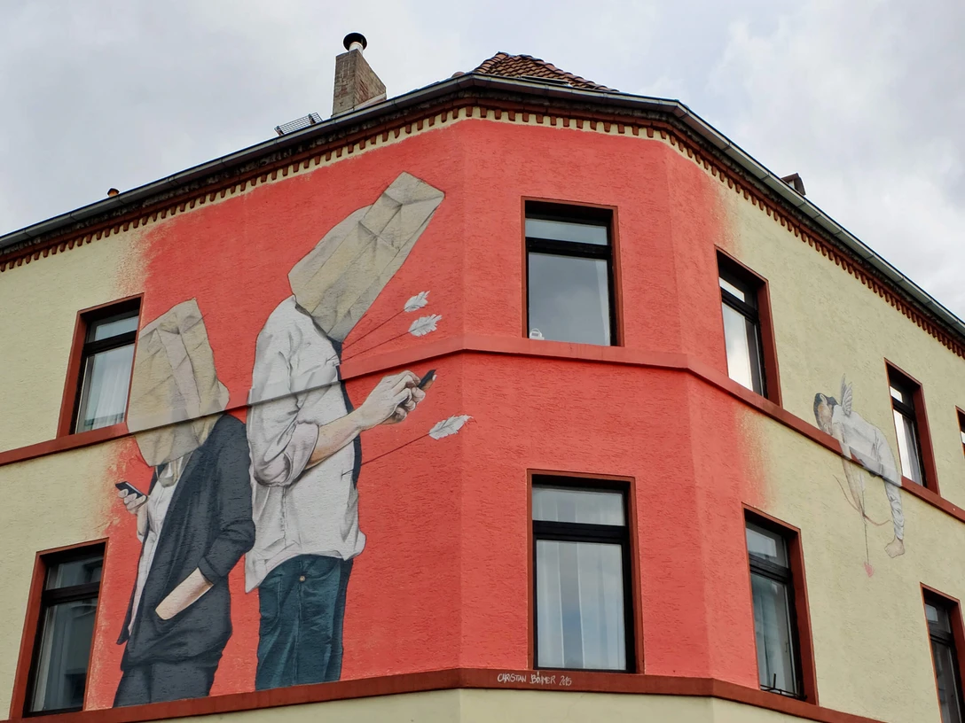 Mural Auf dem Bild ist ein großes Wandgemälde eines Gebäudes an der Ecke einer Straße zu sehen. Zwei Figuren stehen Rücken an Rücken, beide mit Tüten über dem Kopf und Handys in der Hand. Ein Pfeil fliegt von hinten auf die Figuren zu, als ob er von Amor, der links im Bild erscheint, abgeschossen wurde. Die Farben sind hauptsächlich beige und rot, mit schwarzen Fensterumrandungen, die das Gemälde unterbrechen.The picture shows a large mural of a building on the corner of a street. Two figures are standing back to back, both with bags over their heads and cell phones in their hands. An arrow flies towards the figures from behind, as if it has been shot by Cupid, who appears on the left of the picture. The colors are mainly beige and red, with black window borders interrupting the painting.