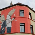 Mural Auf dem Bild ist ein großes Wandgemälde eines Gebäudes an der Ecke einer Straße zu sehen. Zwei Figuren stehen Rücken an Rücken, beide mit Tüten über dem Kopf und Handys in der Hand. Ein Pfeil fliegt von hinten auf die Figuren zu, als ob er von Amor, der links im Bild erscheint, abgeschossen wurde. Die Farben sind hauptsächlich beige und rot, mit schwarzen Fensterumrandungen, die das Gemälde unterbrechen.The picture shows a large mural of a building on the corner of a street. Two figures are standing back to back, both with bags over their heads and cell phones in their hands. An arrow flies towards the figures from behind, as if it has been shot by Cupid, who appears on the left of the picture. The colors are mainly beige and red, with black window borders interrupting the painting.