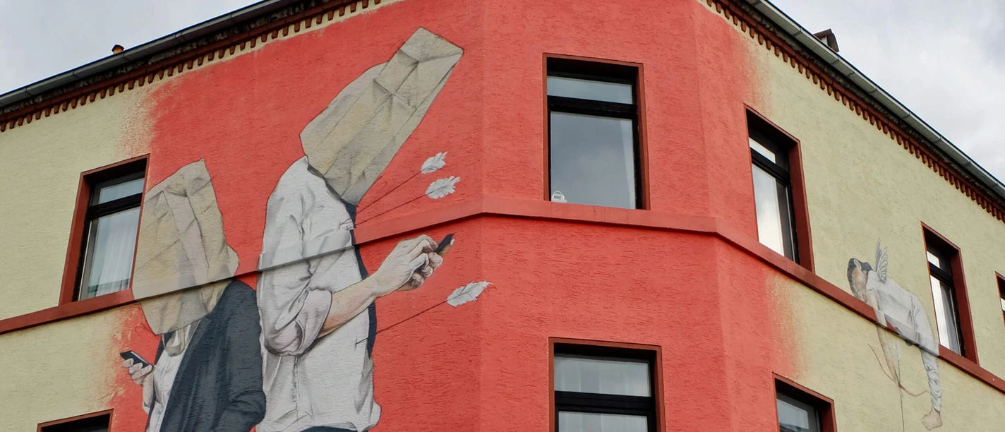 Mural The picture shows a large mural of a building on the corner of a street. Two figures are standing back to back, both with bags over their heads and cell phones in their hands. An arrow flies towards the figures from behind, as if it has been shot by Cupid, who appears on the left of the picture. The colors are mainly beige and red, with black window borders interrupting the painting.