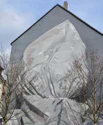 Mural Eine triste, graue Hauswand zeigt ein monumentales Wandgemälde: Ein riesiges, eingewebtes Gesicht, fast transparent und von leichten Falten durchzogen. Vor der Fassade stehen kahle Bäume, die mit ihren Ästen das Bild nicht verdecken, sondern eher umrahmen.