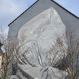 Mural Eine triste, graue Hauswand zeigt ein monumentales Wandgemälde: Ein riesiges, eingewebtes Gesicht, fast transparent und von leichten Falten durchzogen. Vor der Fassade stehen kahle Bäume, die mit ihren Ästen das Bild nicht verdecken, sondern eher umrahmen.A dull, gray house wall shows a monumental mural: a huge, woven-in face, almost transparent and criss-crossed by light folds. Bare trees stand in front of the façade, their branches not obscuring the image, but rather framing it.