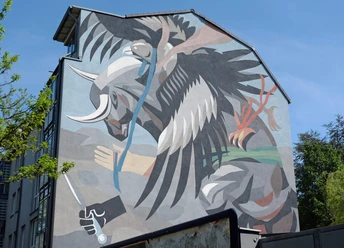 Mural Ein dynamischer Stierkampf zwischen Stier und abstraktem Wesen, in kräftigen Farben an einer Hauswand, von grünen Bäumen eingerahmt.
