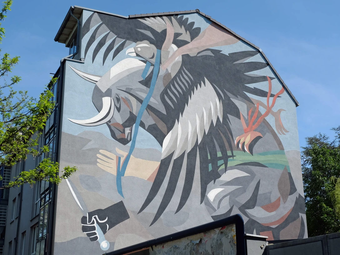 Mural Ein dynamischer Stierkampf zwischen Stier und abstraktem Wesen, in kräftigen Farben an einer Hauswand, von grünen Bäumen eingerahmt.A dynamic bullfight between a bull and an abstract creature, in strong colors on a house wall, framed by green trees.