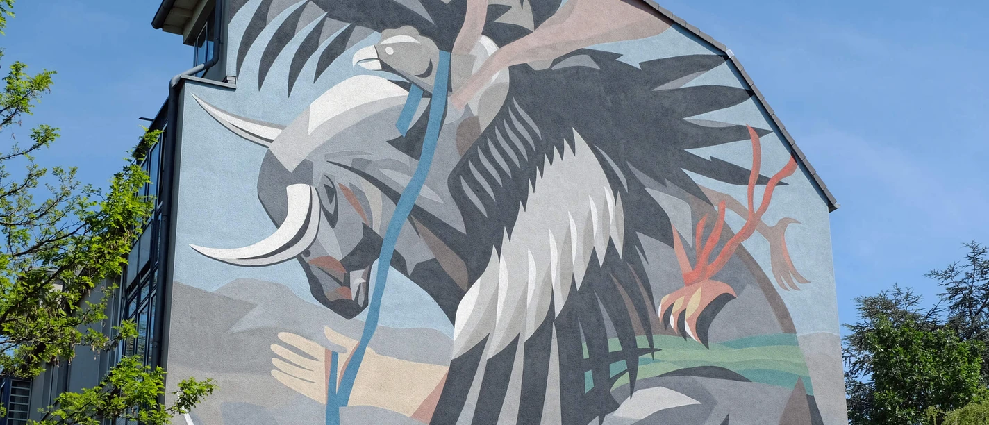 Mural Ein dynamischer Stierkampf zwischen Stier und abstraktem Wesen, in kräftigen Farben an einer Hauswand, von grünen Bäumen eingerahmt.