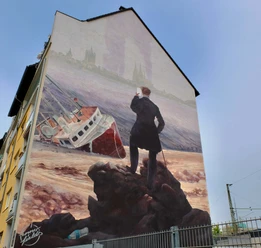 Mural Das Bild zeigt ein Wandgemälde an einem Haus, auf dem ein stilisierter Wanderer steht. Der Wanderer blickt auf ein gestrandetes Schiff vor einer historischen Städtekulisse.
