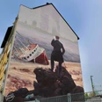 Mural Das Bild zeigt ein Wandgemälde an einem Haus, auf dem ein stilisierter Wanderer steht. Der Wanderer blickt auf ein gestrandetes Schiff vor einer historischen Städtekulisse.The picture shows a mural on a house with a stylized wanderer standing on it. The wanderer is looking at a stranded ship in front of a historic city backdrop.