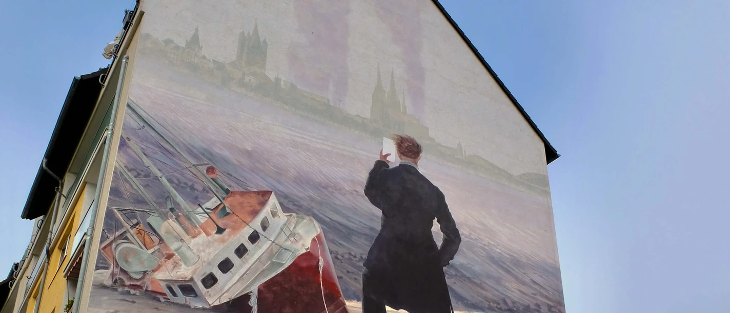 Mural The picture shows a mural on a house with a stylized wanderer standing on it. The wanderer is looking at a stranded ship in front of a historic city backdrop.