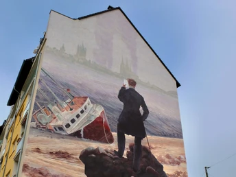 Mural Das Bild zeigt ein Wandgemälde an einem Haus, auf dem ein stilisierter Wanderer steht. Der Wanderer blickt auf ein gestrandetes Schiff vor einer historischen Städtekulisse.The picture shows a mural on a house with a stylized wanderer standing on it. The wanderer is looking at a stranded ship in front of a historic city backdrop.