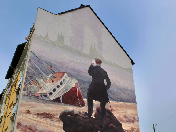 Mural Das Bild zeigt ein Wandgemälde an einem Haus, auf dem ein stilisierter Wanderer steht. Der Wanderer blickt auf ein gestrandetes Schiff vor einer historischen Städtekulisse.