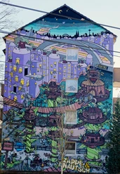 Mural Lebendige Street Art in Köln: Ein Hochhaus mit farbenfrohem Wandgemälde, das eine fantasievolle Stadtlandschaft zeigt, in der überdimensionale Spielfiguren und architektonische Elemente in surrealer Anordnung zu sehen sind. Auffallend sind die dominanten Blau- und Lilatöne sowie die verspielten, märchenhaften Details.