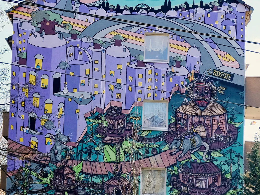 Mural Lebendige Street Art in Köln: Ein Hochhaus mit farbenfrohem Wandgemälde, das eine fantasievolle Stadtlandschaft zeigt, in der überdimensionale Spielfiguren und architektonische Elemente in surrealer Anordnung zu sehen sind. Auffallend sind die dominanten Blau- und Lilatöne sowie die verspielten, märchenhaften Details.Vibrant street art in Cologne: A high-rise building showcasing a colorful mural depicting an imaginative cityscape, featuring oversized game pieces and architectural elements arranged in a surreal manner. Prominent blue and purple tones, along with playful, fairy-tale details, stand out distinctly.