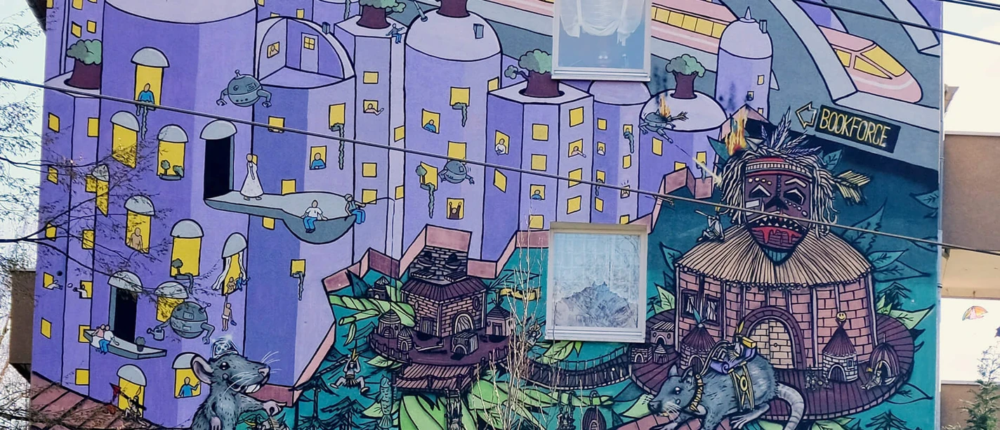 Mural Lebendige Street Art in Köln: Ein Hochhaus mit farbenfrohem Wandgemälde, das eine fantasievolle Stadtlandschaft zeigt, in der überdimensionale Spielfiguren und architektonische Elemente in surrealer Anordnung zu sehen sind. Auffallend sind die dominanten Blau- und Lilatöne sowie die verspielten, märchenhaften Details.