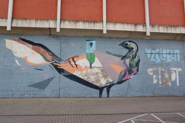 Mural Auf dem Bild ist ein farbenfrohes Graffiti an einer Betonmauer zu sehen. Es zeigt eine künstlerische Darstellung eines Vogels, der in geometrischen Formen und kräftigen Farben gestaltet ist. Oberhalb des Bildes ist eine Backsteinwand zu erkennen, die das Motiv rahmt.