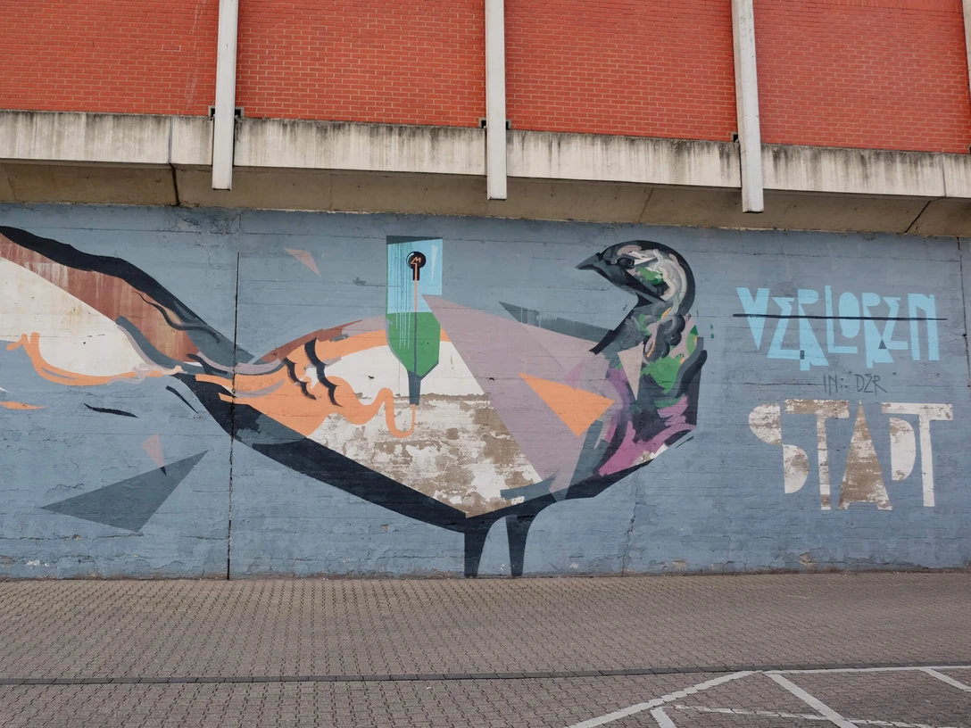 Mural Auf dem Bild ist ein farbenfrohes Graffiti an einer Betonmauer zu sehen. Es zeigt eine künstlerische Darstellung eines Vogels, der in geometrischen Formen und kräftigen Farben gestaltet ist. Oberhalb des Bildes ist eine Backsteinwand zu erkennen, die das Motiv rahmt. The picture shows a colorful graffiti on a concrete wall. It shows an artistic depiction of a bird designed in geometric shapes and bold colors. A brick wall can be seen above the picture, framing the motif.