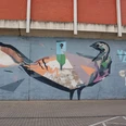 Mural Auf dem Bild ist ein farbenfrohes Graffiti an einer Betonmauer zu sehen. Es zeigt eine künstlerische Darstellung eines Vogels, der in geometrischen Formen und kräftigen Farben gestaltet ist. Oberhalb des Bildes ist eine Backsteinwand zu erkennen, die das Motiv rahmt. The picture shows a colorful graffiti on a concrete wall. It shows an artistic depiction of a bird designed in geometric shapes and bold colors. A brick wall can be seen above the picture, framing the motif.