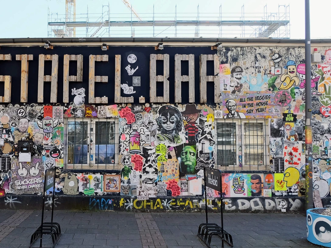 Mural Eine bunte Street-Art-Wand zeigt Graffiti und Sticker in lebhaften Farben, flankiert von Baugerüsten.A colorful street art wall displays graffiti and stickers in vibrant colors, flanked by scaffolding.