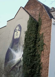 Mural Das Kunstwerk zeigt eine beeindruckende, großflächige Wandmalerei in Köln. Das Bild zeigt eine Madonna mit geschlossenen Augen, goldene Akzente betonen ihr Gesicht. Die Szene wird von üppigem Efeu flankiert, das an der Backsteinmauer hochwächst und einen harmonischen Kontrast zur urbanen Umgebung bildet.
