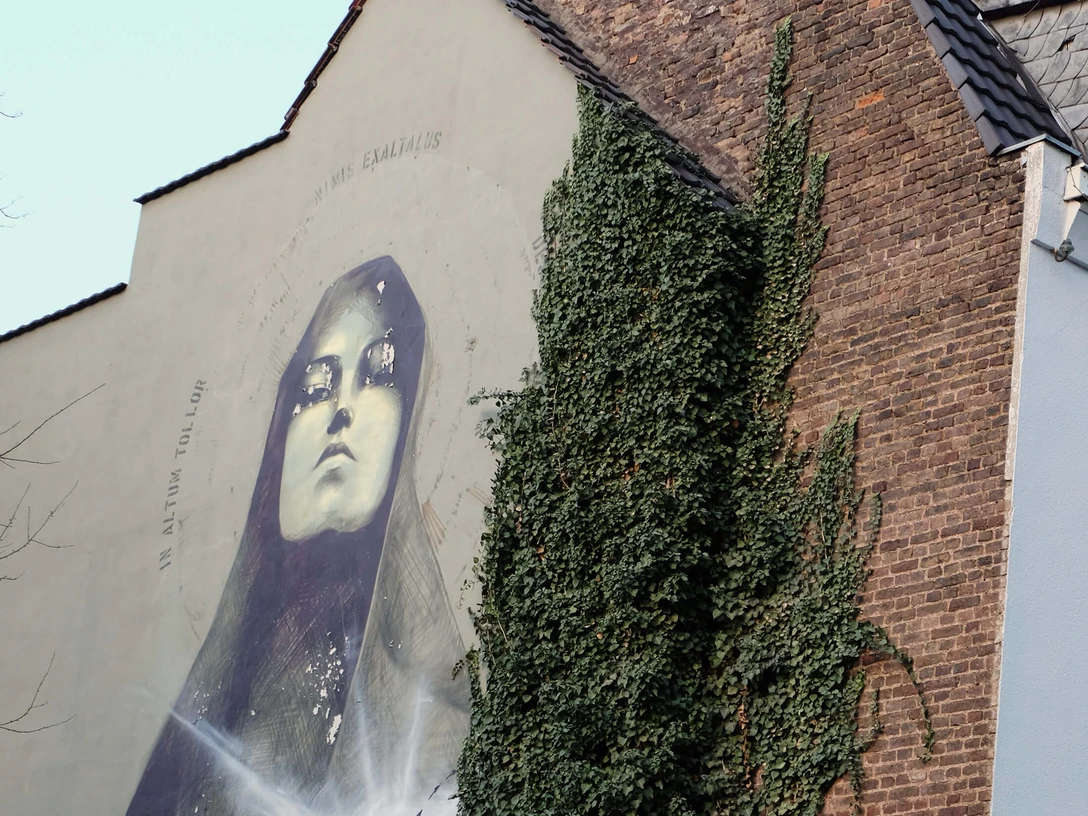 Mural Das Kunstwerk zeigt eine beeindruckende, großflächige Wandmalerei in Köln. Das Bild zeigt eine Madonna mit geschlossenen Augen, goldene Akzente betonen ihr Gesicht. Die Szene wird von üppigem Efeu flankiert, das an der Backsteinmauer hochwächst und einen harmonischen Kontrast zur urbanen Umgebung bildet. The artwork shows an impressive, large-scale mural in Cologne. The picture shows a Madonna with closed eyes, golden accents emphasize her face. The scene is flanked by lush ivy growing up the brick wall, creating a harmonious contrast to the urban surroundings.