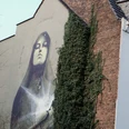 Mural Das Kunstwerk zeigt eine beeindruckende, großflächige Wandmalerei in Köln. Das Bild zeigt eine Madonna mit geschlossenen Augen, goldene Akzente betonen ihr Gesicht. Die Szene wird von üppigem Efeu flankiert, das an der Backsteinmauer hochwächst und einen harmonischen Kontrast zur urbanen Umgebung bildet. The artwork shows an impressive, large-scale mural in Cologne. The picture shows a Madonna with closed eyes, golden accents emphasize her face. The scene is flanked by lush ivy growing up the brick wall, creating a harmonious contrast to the urban surroundings.