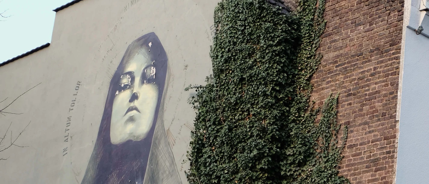 Mural The artwork shows an impressive, large-scale mural in Cologne. The picture shows a Madonna with closed eyes, golden accents emphasize her face. The scene is flanked by lush ivy growing up the brick wall, creating a harmonious contrast to the urban surroundings.