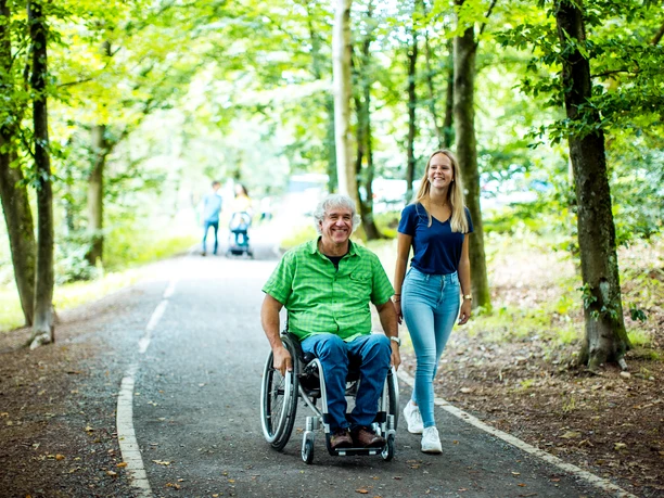 barrierefreier Weg an der Brucher Talsperre Senior im Rollstuhl auf Waldweg, begleitet von junger Frau; beide lächeln in grüner Umgebung.