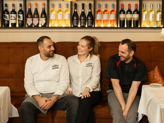 (C) La Fucina 1.jpg Drei Restaurantmitarbeiter in Uniform sitzen lächelnd auf einer Bank vor einer gefüllten Weinregalwand.