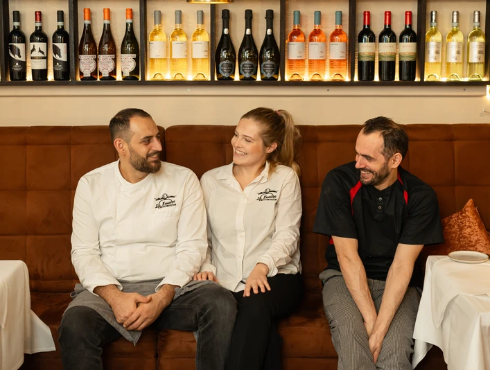 (C) La Fucina 1.jpg Drei Restaurantmitarbeiter in Uniform sitzen lächelnd auf einer Bank vor einer gefüllten Weinregalwand.