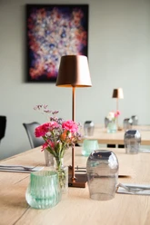 Tisch mit Gläsern, Teelicht, Vase mit Blumen, Besteck, Servietten und einer kleinen Stehlampe. Im Hintergrund hängt ein Bild an der Wand.